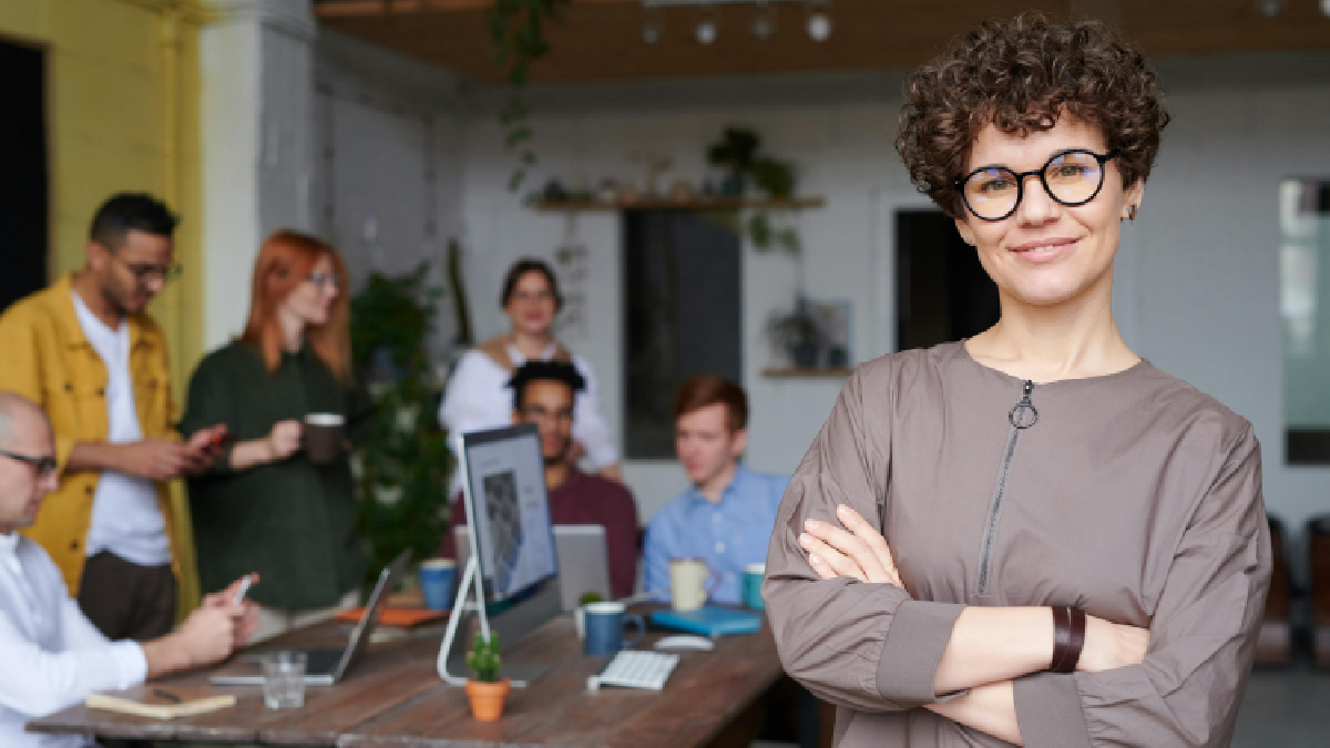 Mulher de braços cruzados em posição de liderança a frente a mesa de pessoas trabalhando. Foto: Pexels
